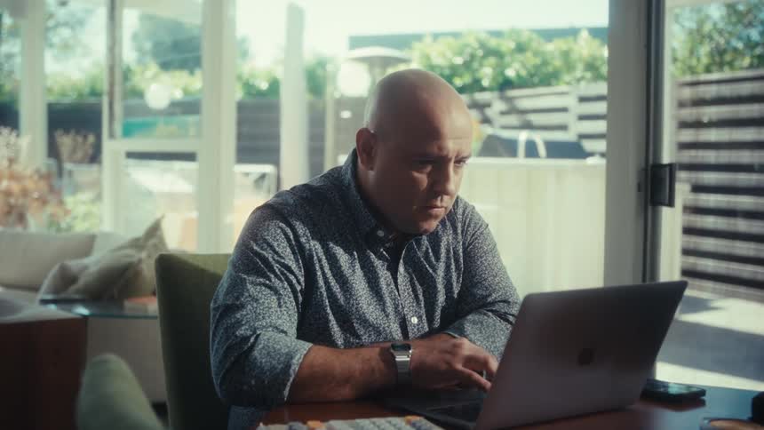 Man working on laptop at table with notebook and pen in room with large glass windows