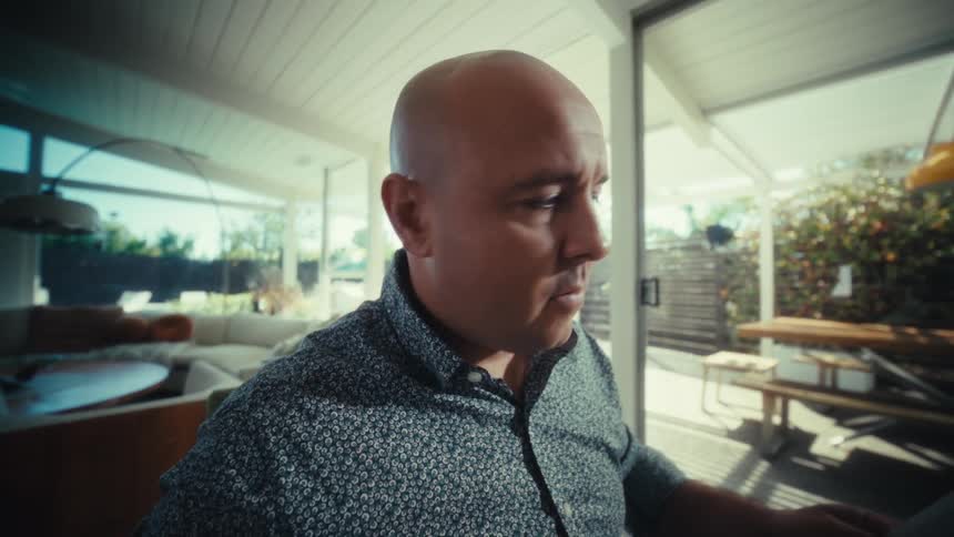 A bald man sits at a wooden table in a bright room looking at printed bar charts while using a calculator and typing on a laptop