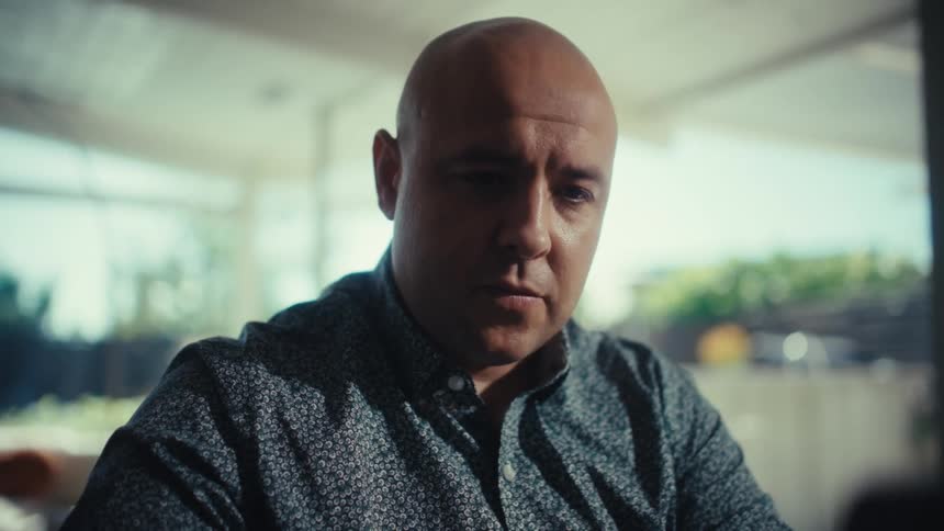 A bald man in a patterned shirt sits indoors looking down with a serious and pensive expression