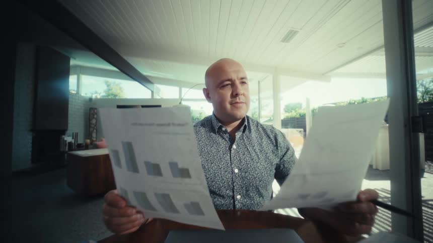 Man seated at a table reviews two sheets with charts beside a laptop and calculator