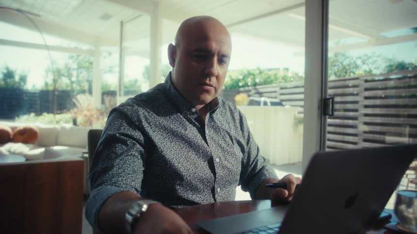 Man sitting at table using laptop with glass doors and outdoor area in the background