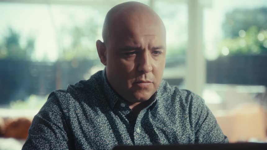 A bald man in a patterned shirt works intently on a laptop at a table zooming in on his face