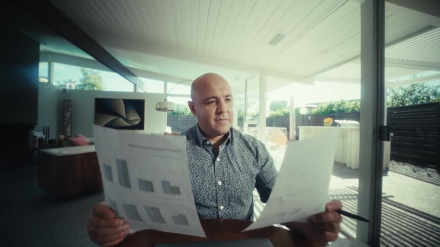 A man reviews financial documents with charts and a calculator at a table in a modern home then smiles and nods