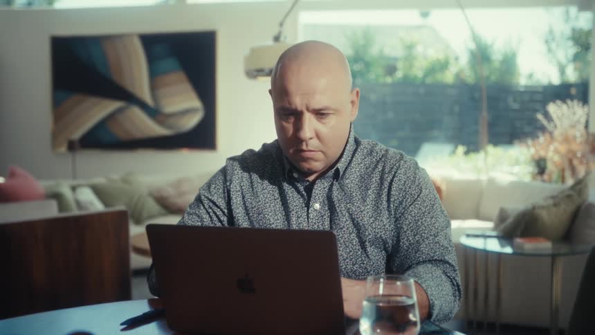 A bald man works on a laptop at a wooden table in a modern living room with large windows