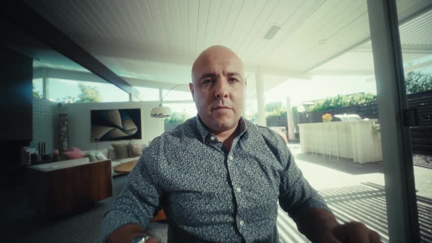The video shows a bald man in a patterned blue shirt sitting at a table in a bright modern room he begins by looking at a laptop and then leans in closer toward the camera with a focused expression