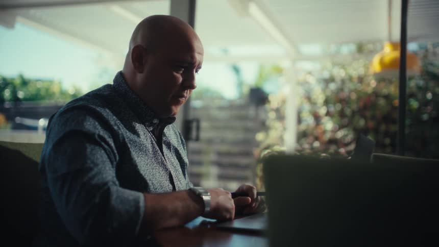 A bald man in a patterned shirt sits at a table focused on his laptop while holding a penthe camera slowly pans out to reveal a glass of water on the table and a bright outdoor patio in the background