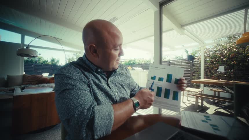A man is sitting at a wooden table in a brightly lit modern home and looking at a laptop he then picks up a sheet of paper with bar graphs on it and begins pointing at the data with a pen