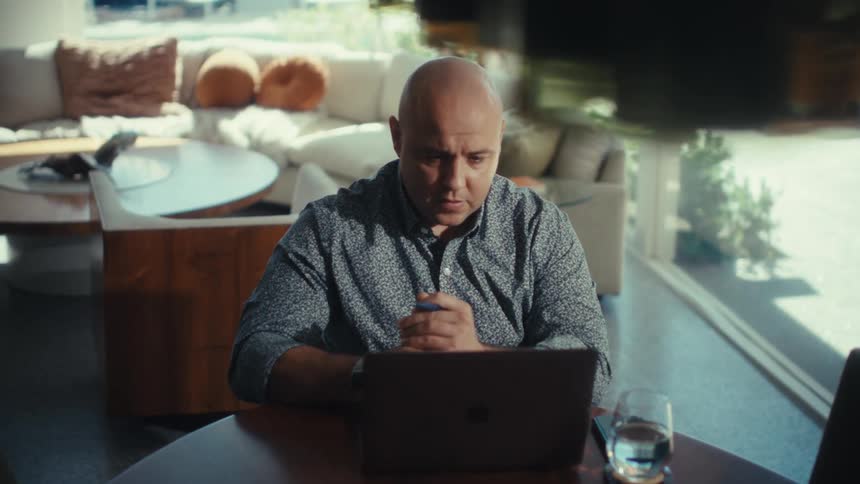 A bald man in a patterned blue shirt sits at a round wooden table indoors and looks at his laptop screen with a serious expression while holding a pen near a glass of water with a bright living room in the background