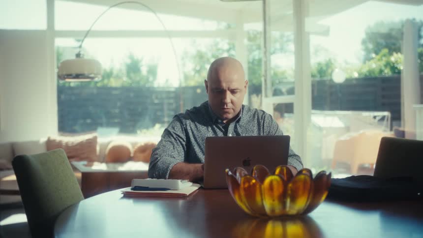 A bald man works on a laptop at a table in a bright modern home