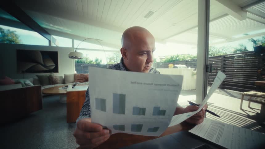 Man sitting at a table in front of a laptop computer as he looks at paperwork and presses keys on a calculator