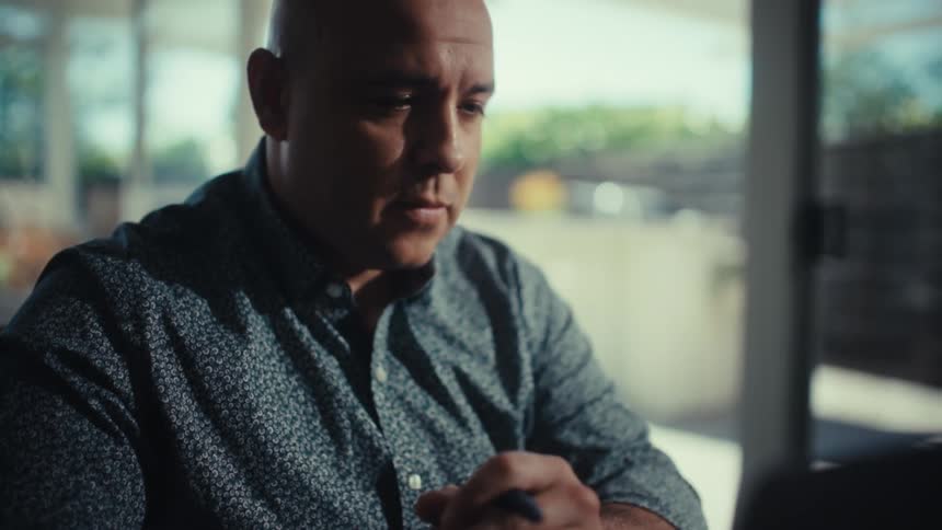 A man sits at a desk with his hands clasped staring intently at a laptop screen in a brightly lit roomthe camera slowly tilts up from his hands and watch to focus on his serious facial expression as he works