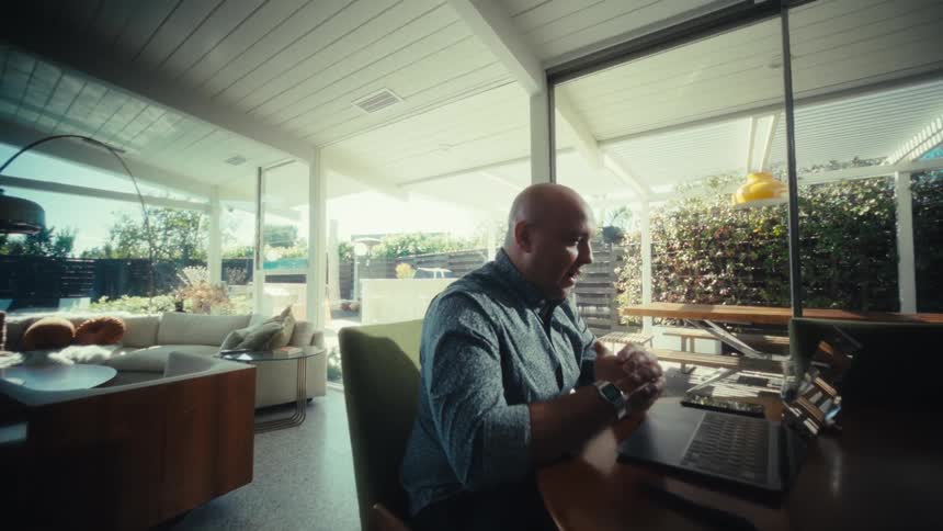 A bald man in a patterned shirt sits at a wooden dining table and talks during a video call on his laptop inside a bright modern home with large glass doors and a view of the patio