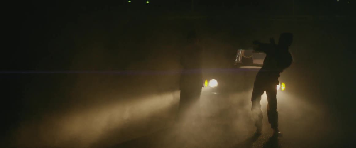 People dancing outdoors around flashing lights and a car's headlights shining behind them