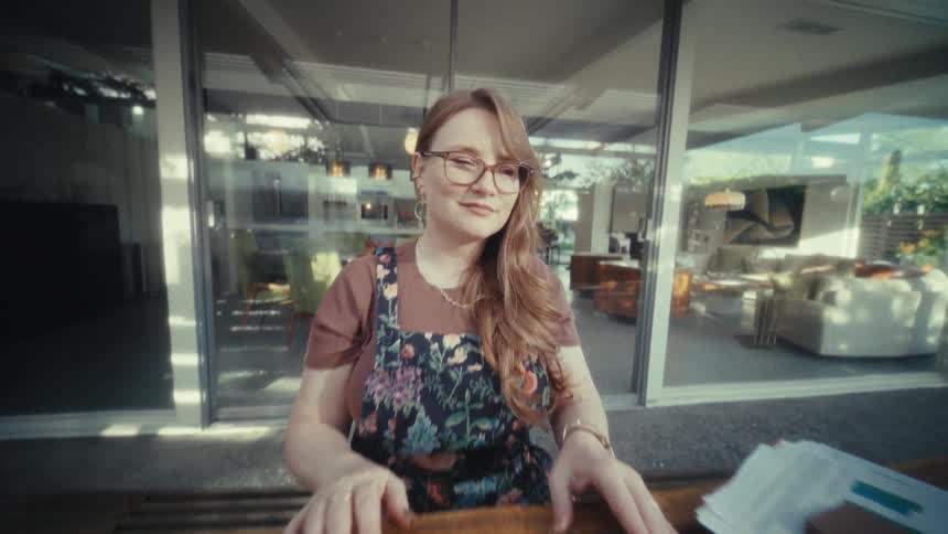 A woman closes her laptop and smiles while sitting at a wooden table on a patio