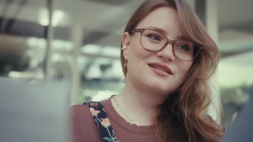 A smiling redhead woman with glasses and floral overalls talks to someone offcamera while holding papers in a bright indoor setting
