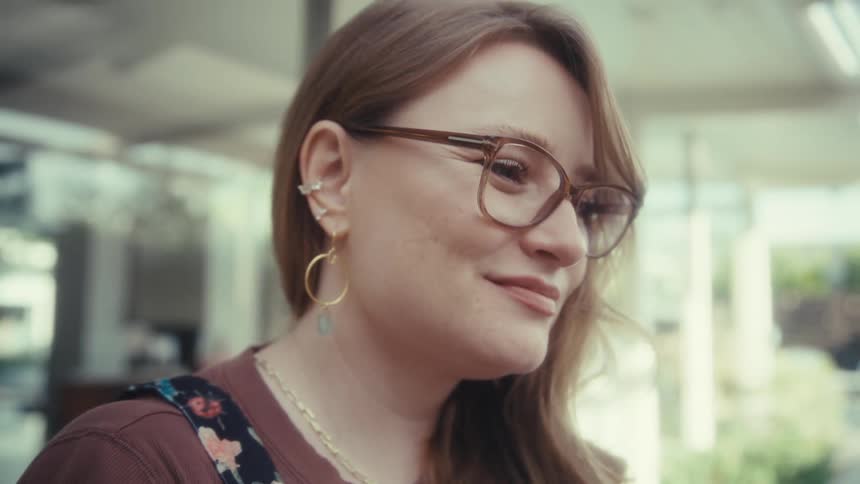 A young woman wearing glasses sits outdoors smiling as she types on a computer