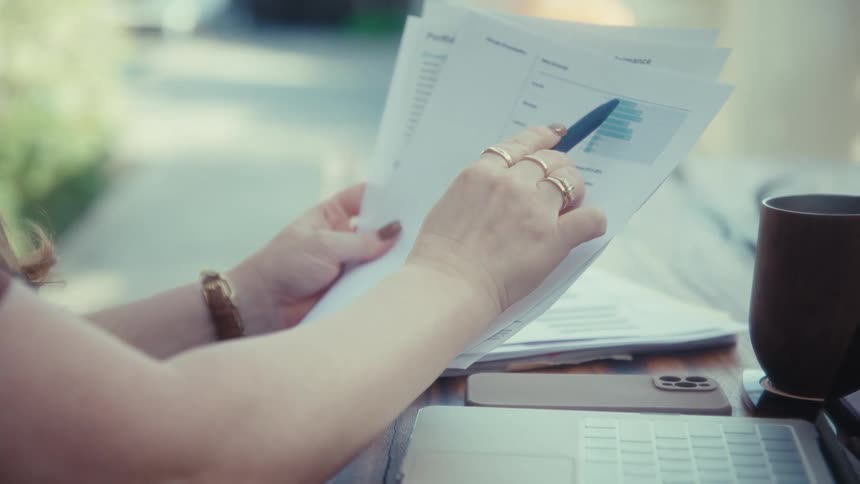 A person reviewing printed documents at an outdoor table using a pen to point at specific datathe first line of interest is a blue pen held in the persons right hand which they use to gesture toward various charts and figures on the pages
