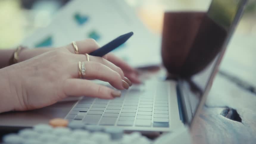 A woman with gold rings and brown nails types on a laptop at a wooden table while holding a blue pen and looking at printed bar charts in a bright outdoor setting
