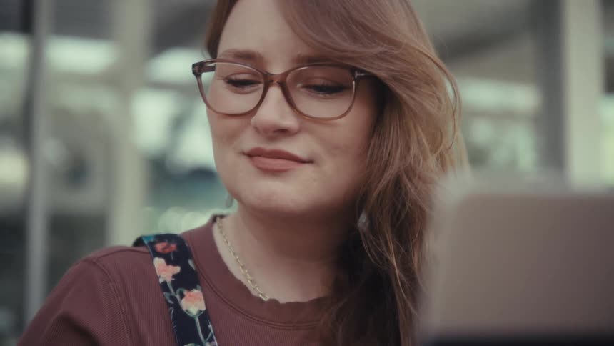 A woman with glasses sits at a wooden table and uses a laptop
