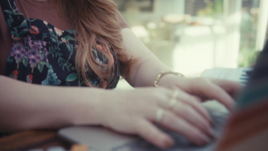 The video features a woman in glasses and floral overalls smiling while sitting at an outdoor table the camera then pans down to show her hands typing on a laptop with colorful stripes on the back positioned next to a calculator
