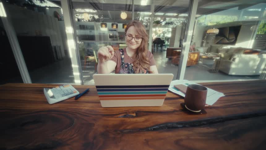 A woman wearing glasses and overalls sits at a large wooden desk typing on a laptop decorated with colorful horizontal stripes