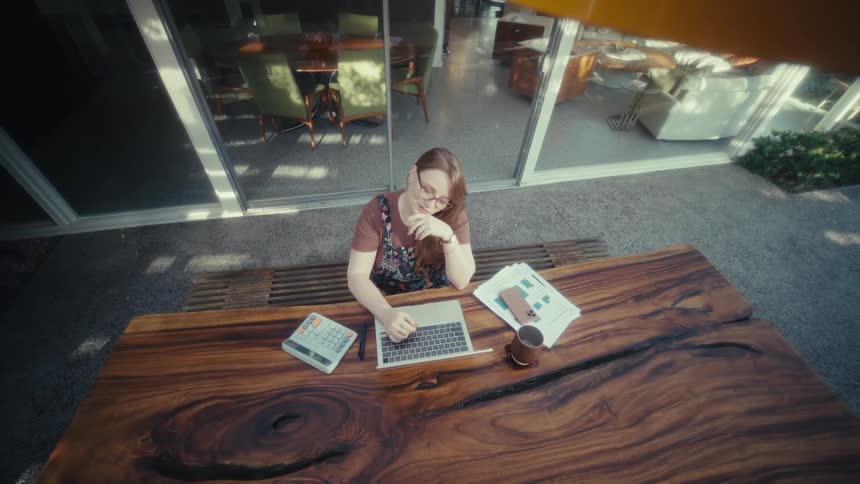 Aerial view of woman working on laptop at wooden table with calculator reports and mug beside her