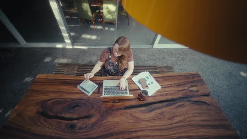 A young woman wearing glasses sits outdoors on a bench at midday typing on her computer at a desk in front of her