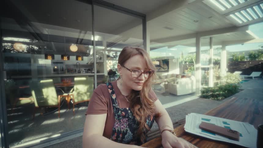 In this video a woman wearing glasses is sitting at a large wooden outdoor table and opens her laptop to begin working there is a calculator and some paperwork next to her as she starts typing with a smile