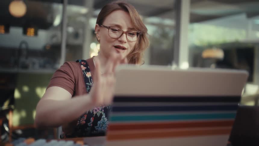 A young woman wearing glasses sits at a dining table at midday typing on a computer keyboard