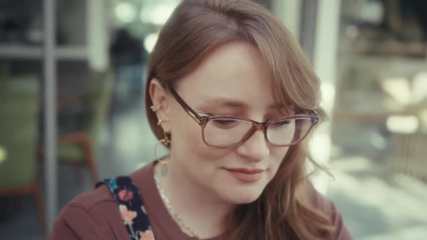 A young woman wearing glasses sits outdoors at midday sitting quietly in deep thought with a smile on her face