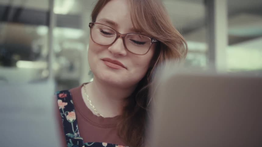 A woman wearing glasses and a floral top holds a paper with a bar graph while looking at a laptop screen and then makes a doubtful facial expression while sitting at a wooden table outdoors