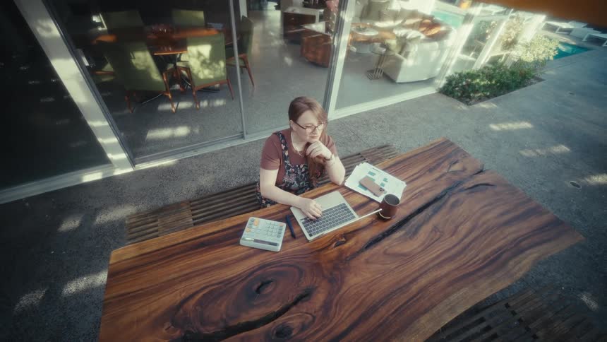 A woman wearing glasses sits on a bench outside an office building at midday typing on a laptop