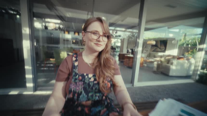 A woman in glasses finishes typing on her laptop closes the lid and leans back with a satisfied smile she appears relieved and content as she completes her work at a wooden table