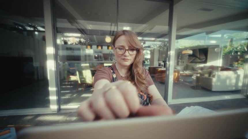 A young woman wearing glasses sits outdoors opens her laptop and starts typing with a smile on her face