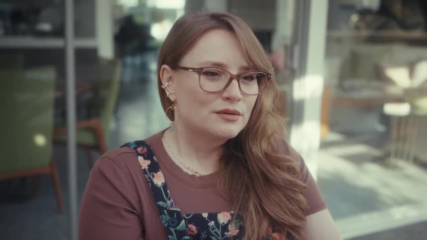 A woman with long auburn hair and glasses sits outside a cafe wearing a brown shirt and floral overallsshe maintains a thoughtful expression as she looks off to the side occasionally nodding slightly while listening or observing
