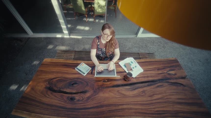 A woman sitting at a wooden table uses a calculator and a laptop
