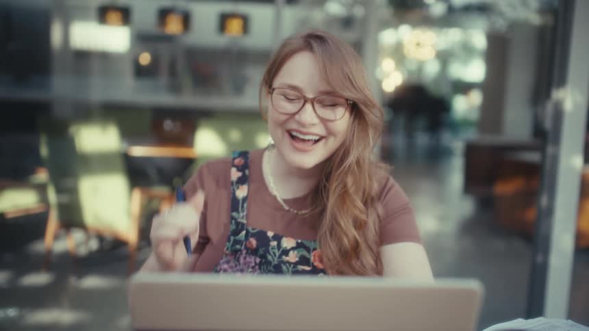 A woman wearing glasses and a floral jumpsuit sits at a table looking at her laptop with a shocked and joyous expressionshe pumps her fist in the air and laughs excitedly appearing to celebrate good news or a major success