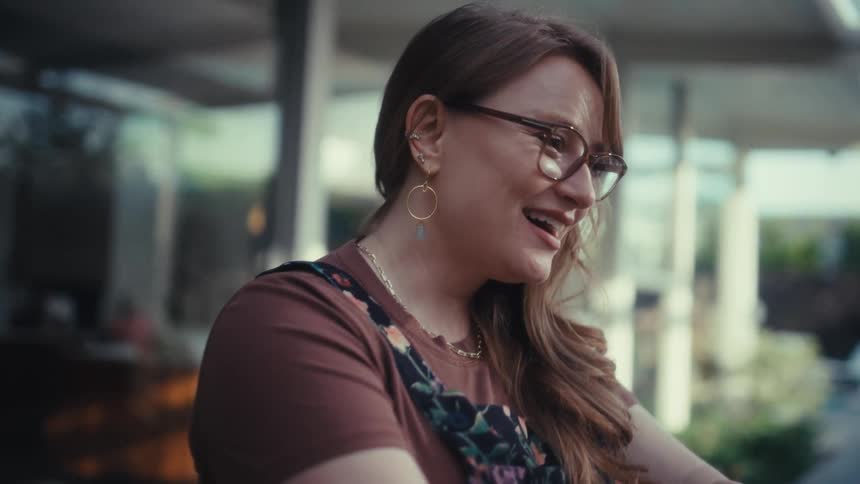A woman wearing glasses and a floral dress sits at a wooden table outside a modern house in front of an open laptop during a bright day