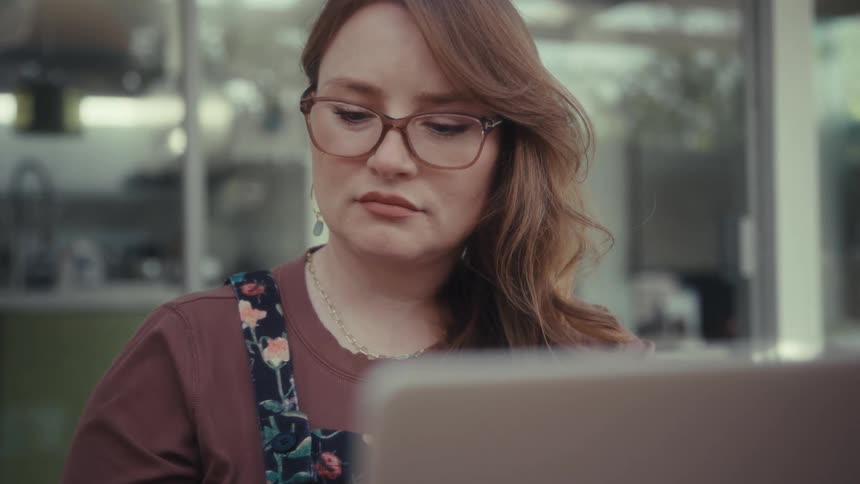 A woman wearing glasses and a floral patterned top sits at a wooden table outdoors and looks at her laptop screen with a focused and then a slightly smiling expression during a bright day