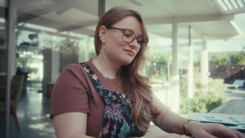 A woman with glasses and long hair sits at a wooden table on a bright patio while typing on her laptop and smiling softly
