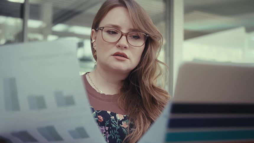 Woman wearing glasses reviewing charts on sheets of paper beside stripedcover laptop