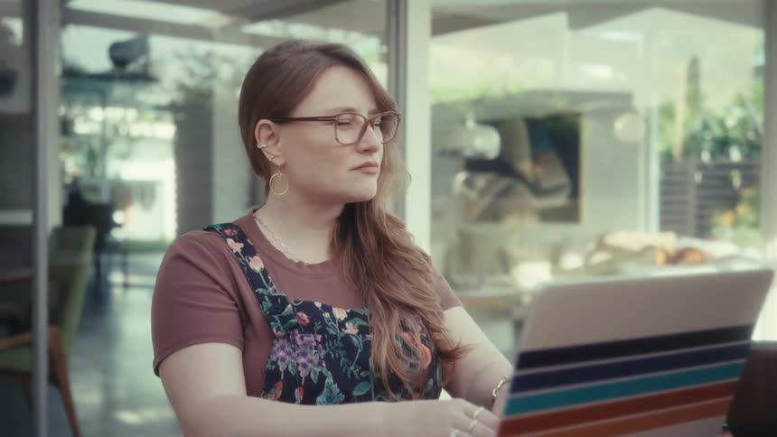 A young woman sits outdoors typing on a computer pausing for a moment in deep thought with a smile before continuing