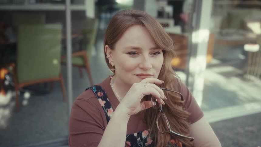 A young woman wearing glasses sits outdoors at midday sitting quietly in deep thought with a smile on her face