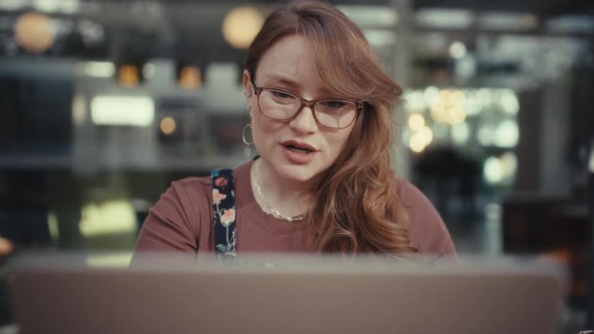 A woman wearing glasses and a floral top sits in front of an open laptop while talking and laughing during a video call at a wooden table in a bright modern setting