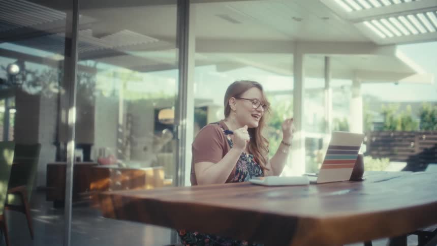 A woman wearing glasses and a floral dress sits at a wooden table outside a modern house while cheering and dancing with joy in front of an open laptop during a bright day
