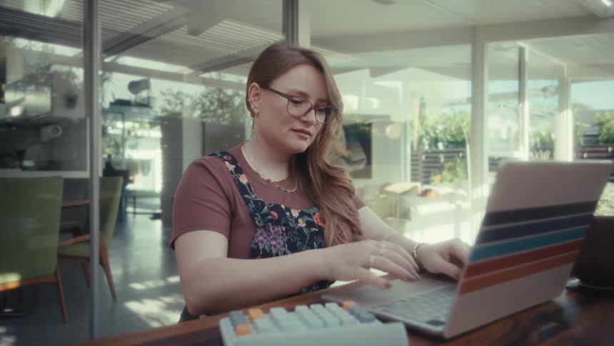 A woman with glasses and long hair working on a laptop at a large wooden table in a sunlit room she appears focused as she types with a colorful keyboard and a mug nearby in the bright modern setting