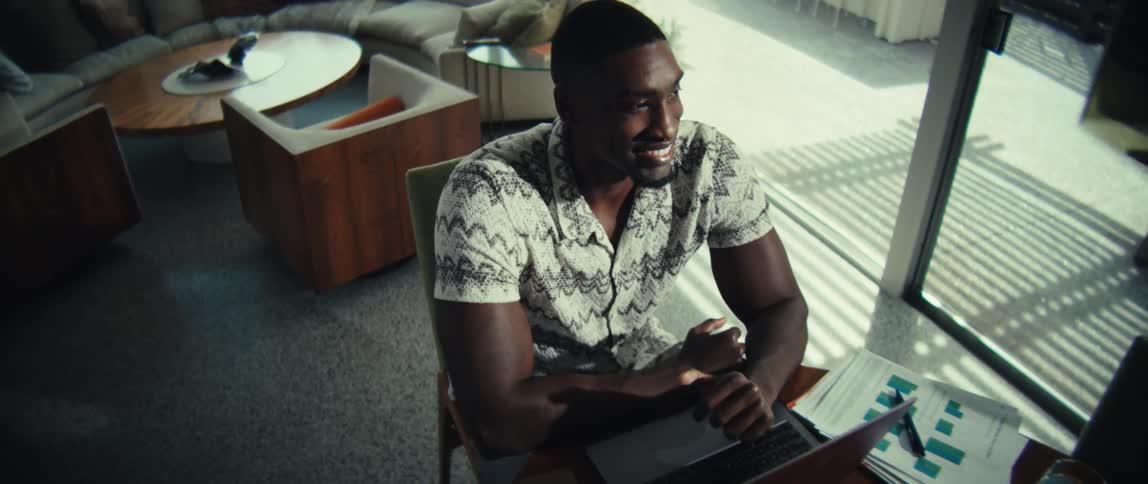 A man sits at a wooden table in a bright modern room while working on a laptop after typing for a moment he pauses to look away from the screen with a satisfied smile