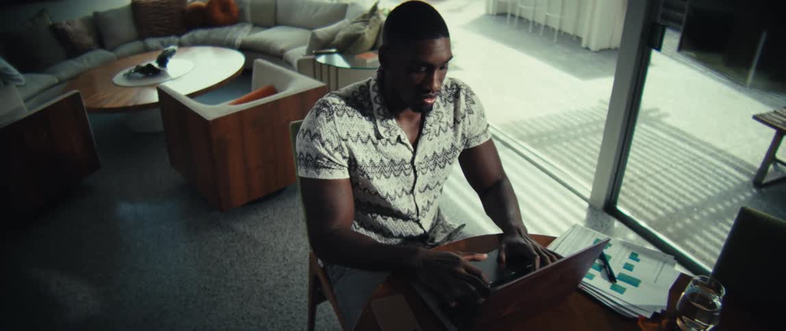 A man sitting at a wooden table in a spacious modern room while typing on a laptop with a woodgrain finish he is surrounded by documents a calculator and a glass of water as he focuses on his work near a large window