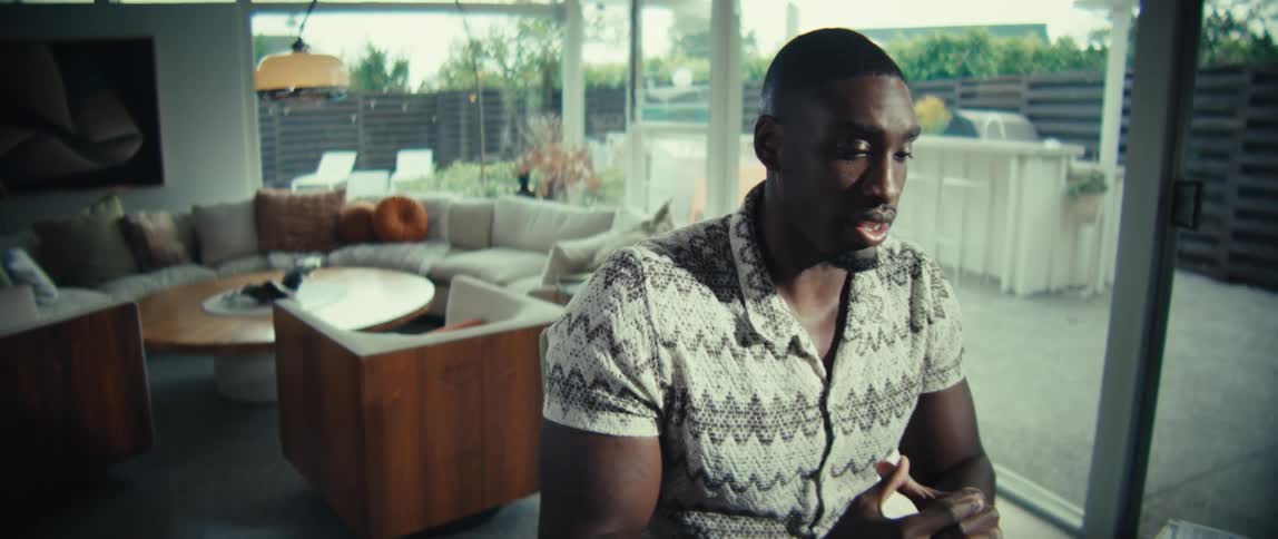 A black man in a patterned shirt sits at a wooden table and speaks while looking at a laptop