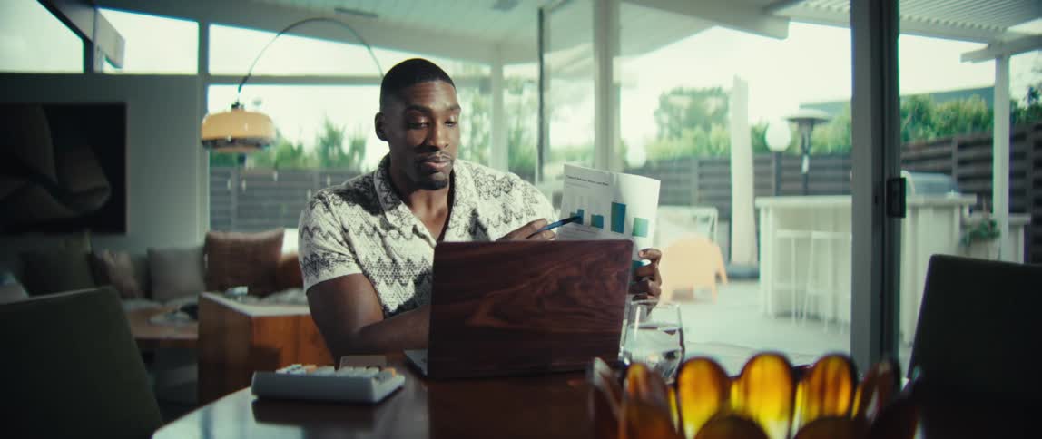 A man wearing a patterned short sleeve shirt sits at a wooden table and holds up a document with green bar charts while pointing and speaking during a video call on a laptop in a bright modern home
